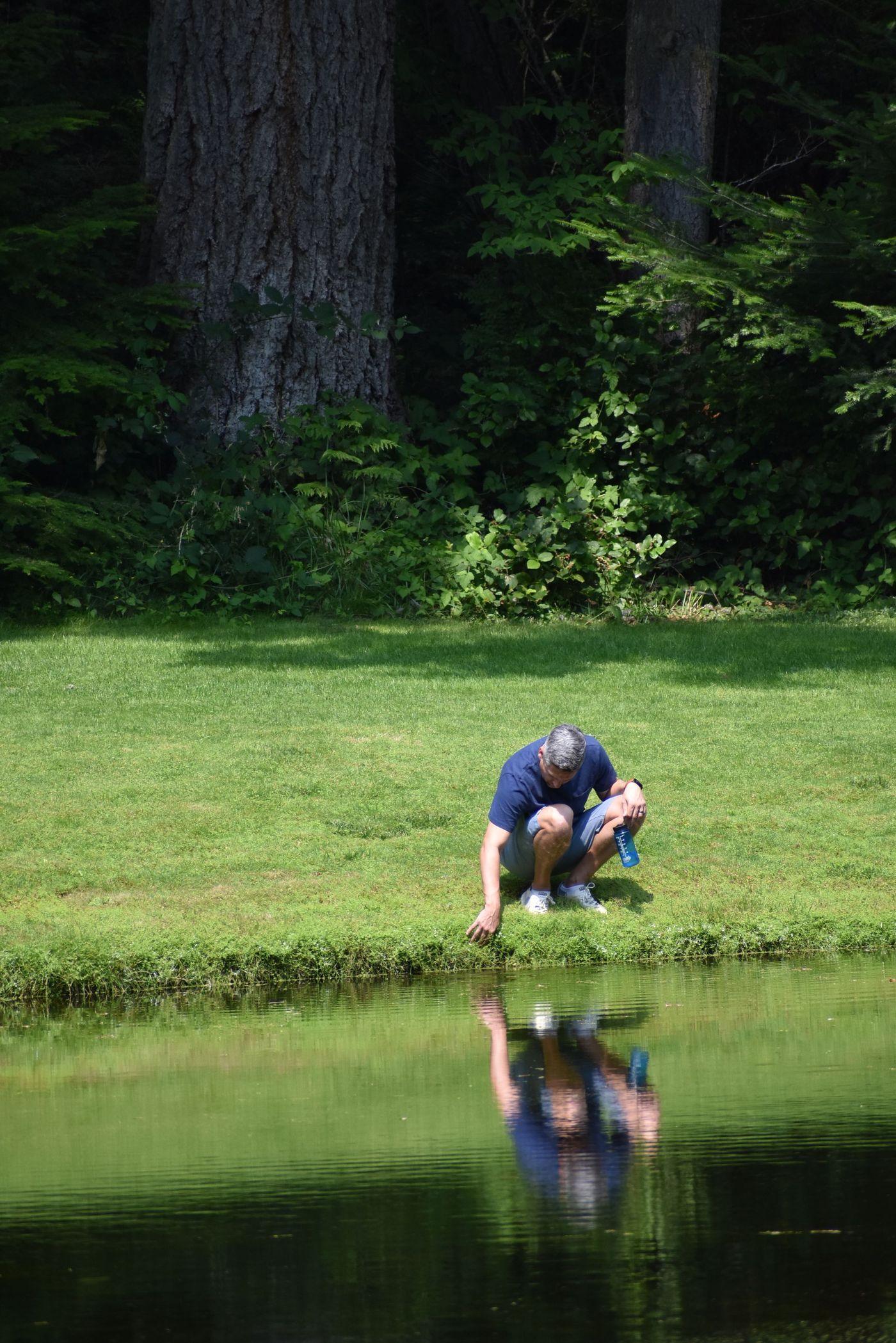 A man crouching at the edge of a pond, examining the water by hand